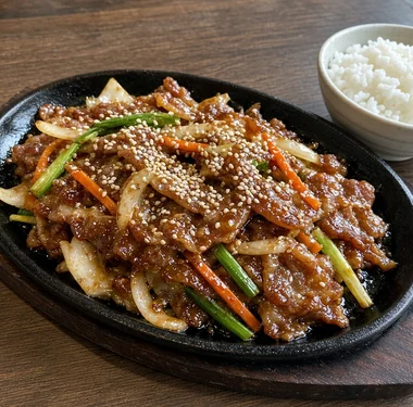 Beef bulgogi with onion, green onion, carrot and sesame seeds on a black sizzling platter, with a bowl of rice