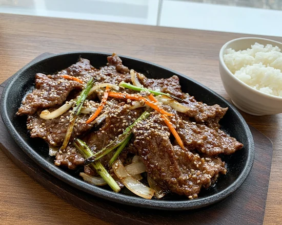 Thin slices of beef bulgogi with onion, green onion and sesame seeds on a black sizzling platter, with a bowl of rice